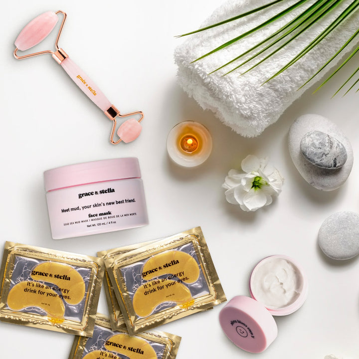 Flat lay of skincare products with a jade roller, towels, stones, candle, eye cream from grace & stella's energizing set and a plant on a white background.